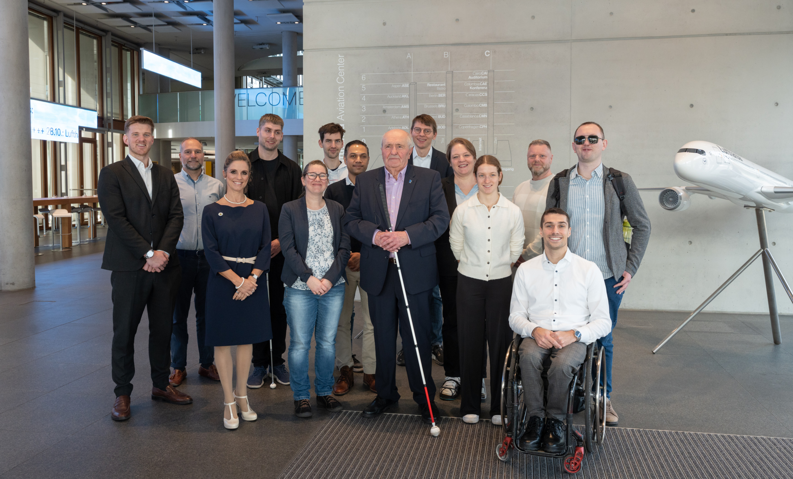 Gruppenfoto des Barrierefreiheits-Beirats der Lufthansa Group im Lufthansa Aviation Center, darunter ein Mann mit weißem Langstock und ein Mann im Rollstuhl, rechts ein Flugzeugmodell.