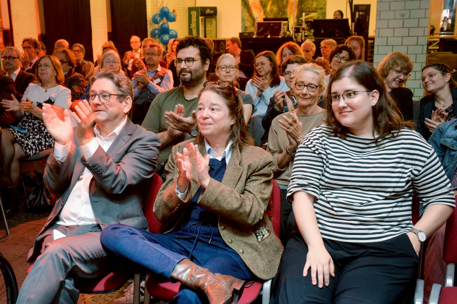 Gäste der Jubiläumsfeier sitzen im Saal und applaudieren.