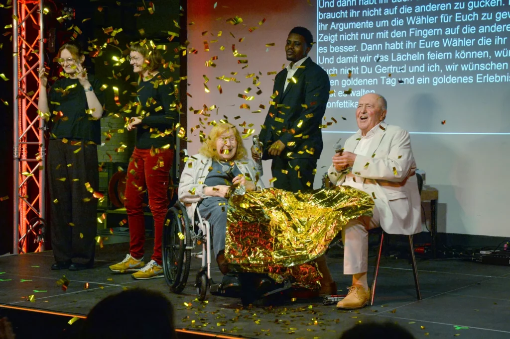 Gertrud Servos und Peter Gabor auf der Bühne der Jubiläumsfeier „30+1 Jahre Landesbehindertenrat NRW“, goldenes Konfetti regnet herab.